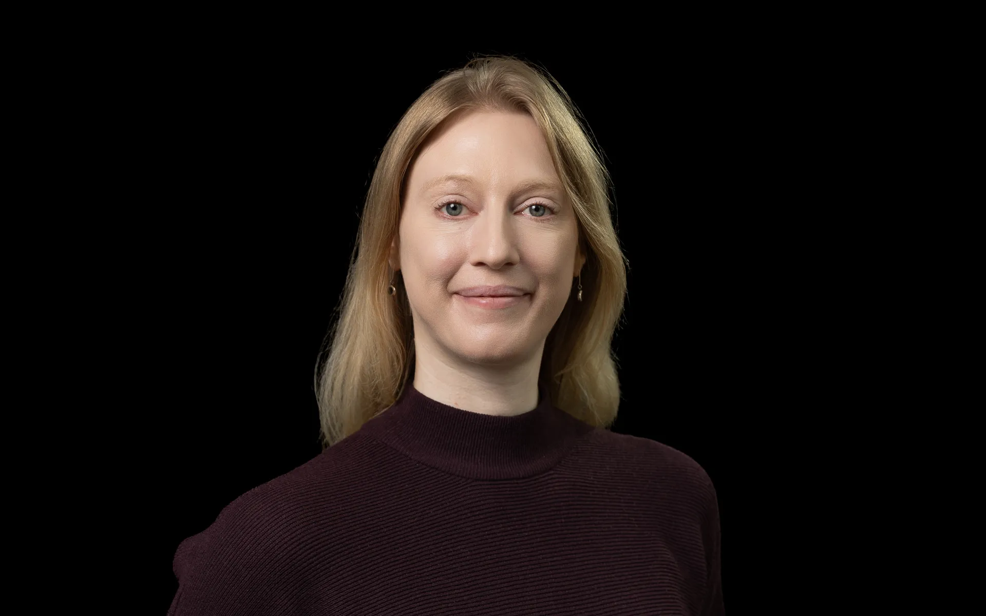 Portrait photo of Sarah smiling at the camera, wearing a dark polo neck top.