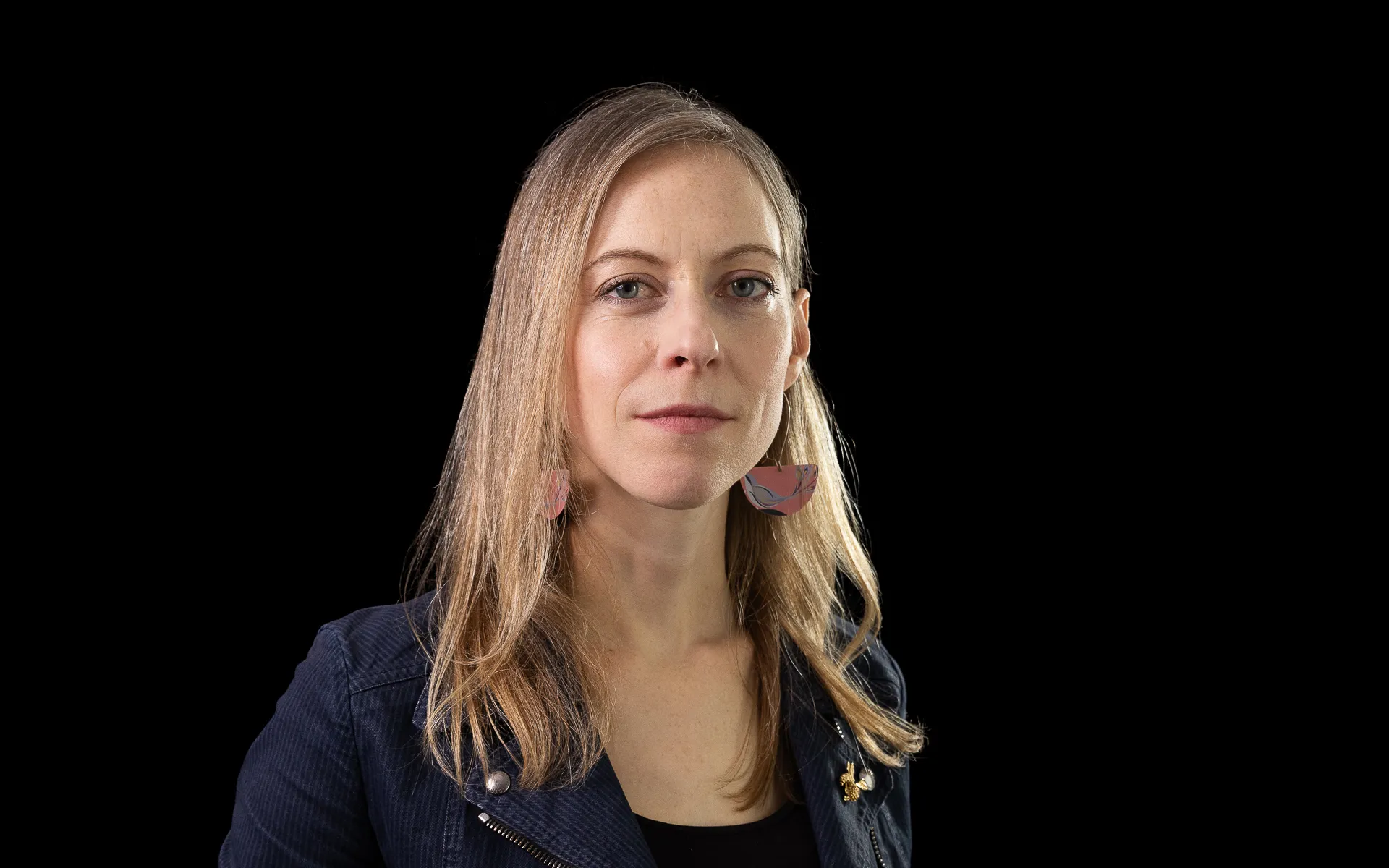 Studio portrait of a woman with long blonde hair wearing a dark jacket over a top, photographed against a black background.