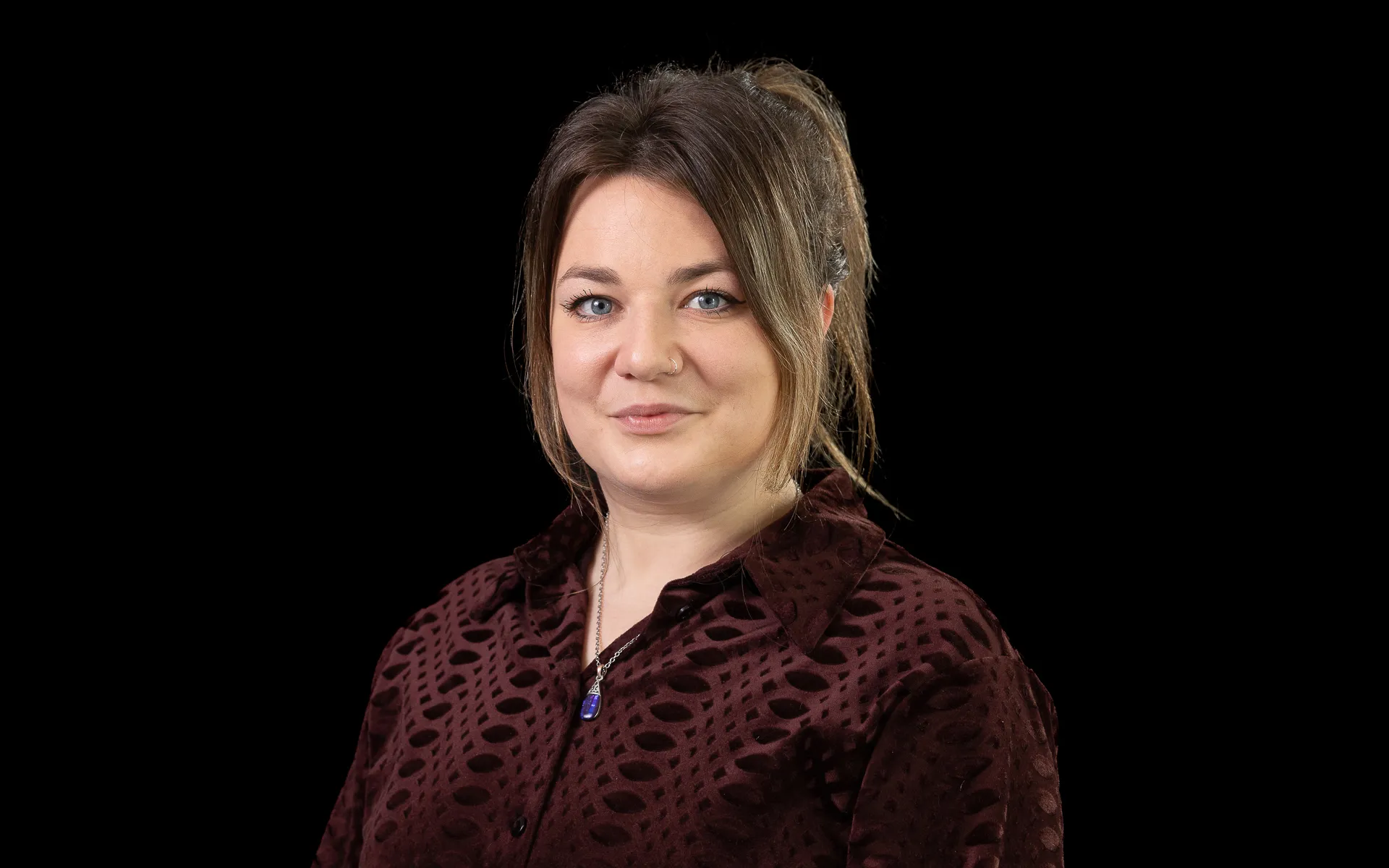 Studio portrait of a woman with hair tied back, wearing a dark patterned blouse and a necklace, photographed against a black background.