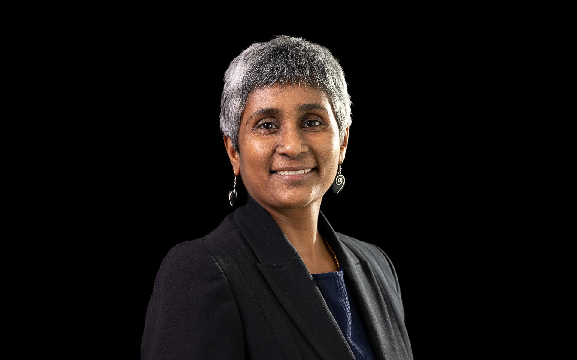 Studio portrait of a woman with short grey hair wearing a dark blazer over a top, photographed against a black background.