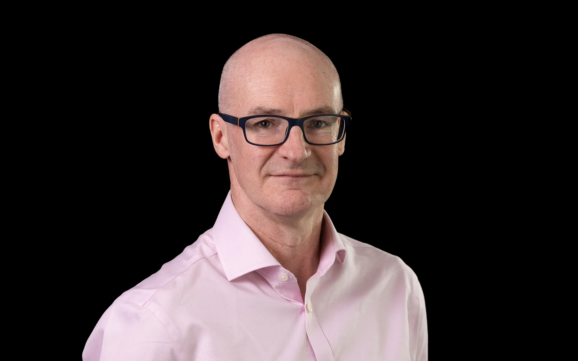 Studio portrait of a man wearing glasses and a light pink shirt, photographed against a black background.