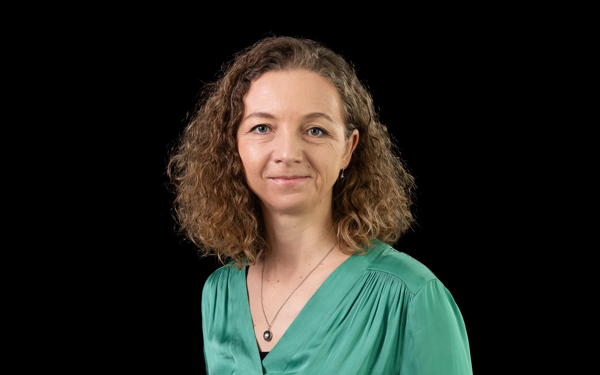 Studio portrait of a woman with curly shoulder-length hair wearing a green blouse and necklace, photographed against a black background.