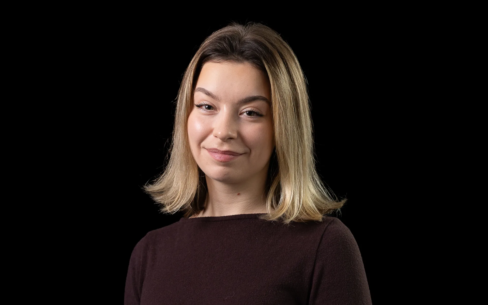 Studio portrait of a woman with shoulder-length blonde hair wearing a dark long-sleeved top, photographed against a black background.