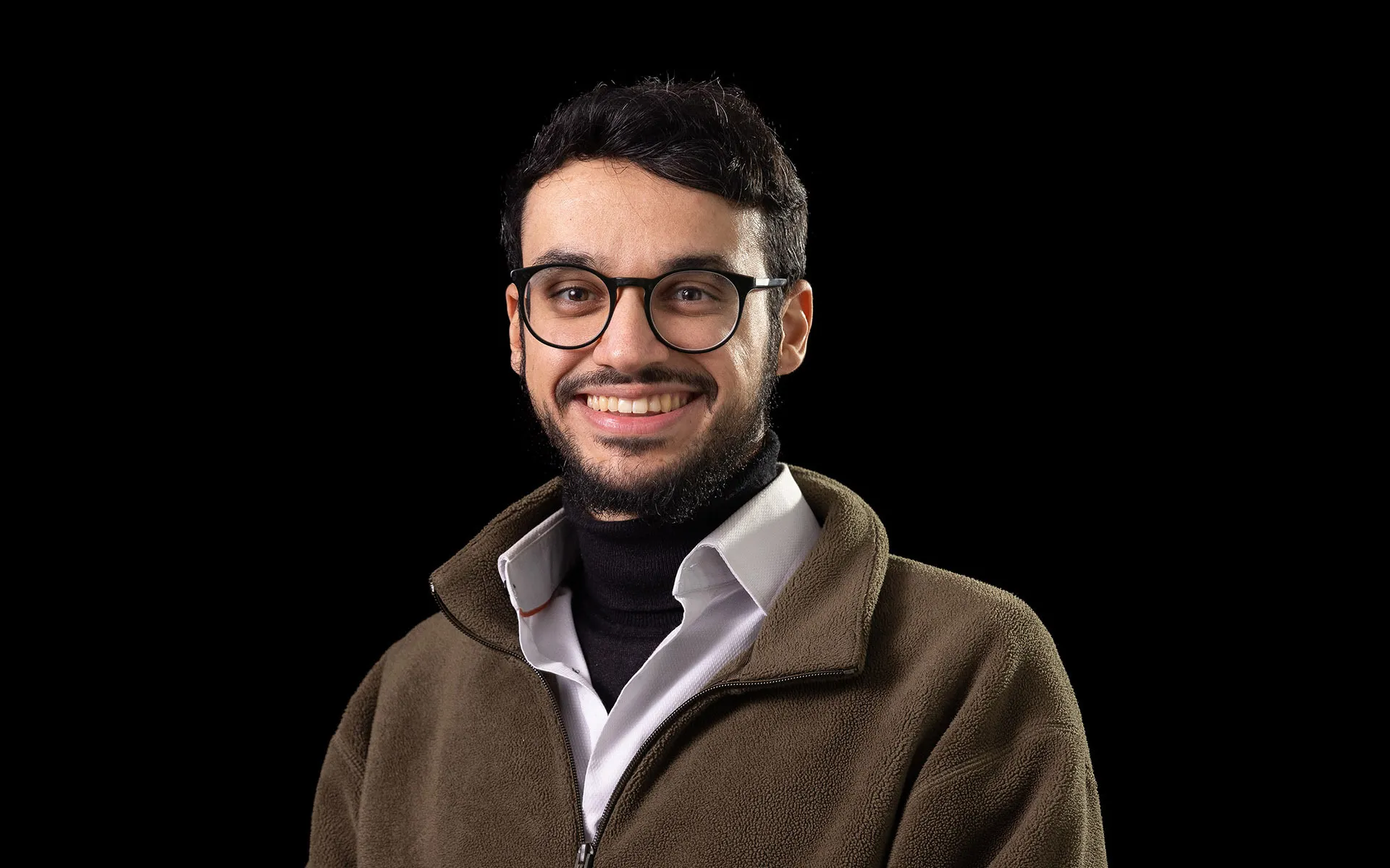 Studio portrait of a person wearing a white collared shirt layered under a dark zip-up fleece, standing against a plain black background.