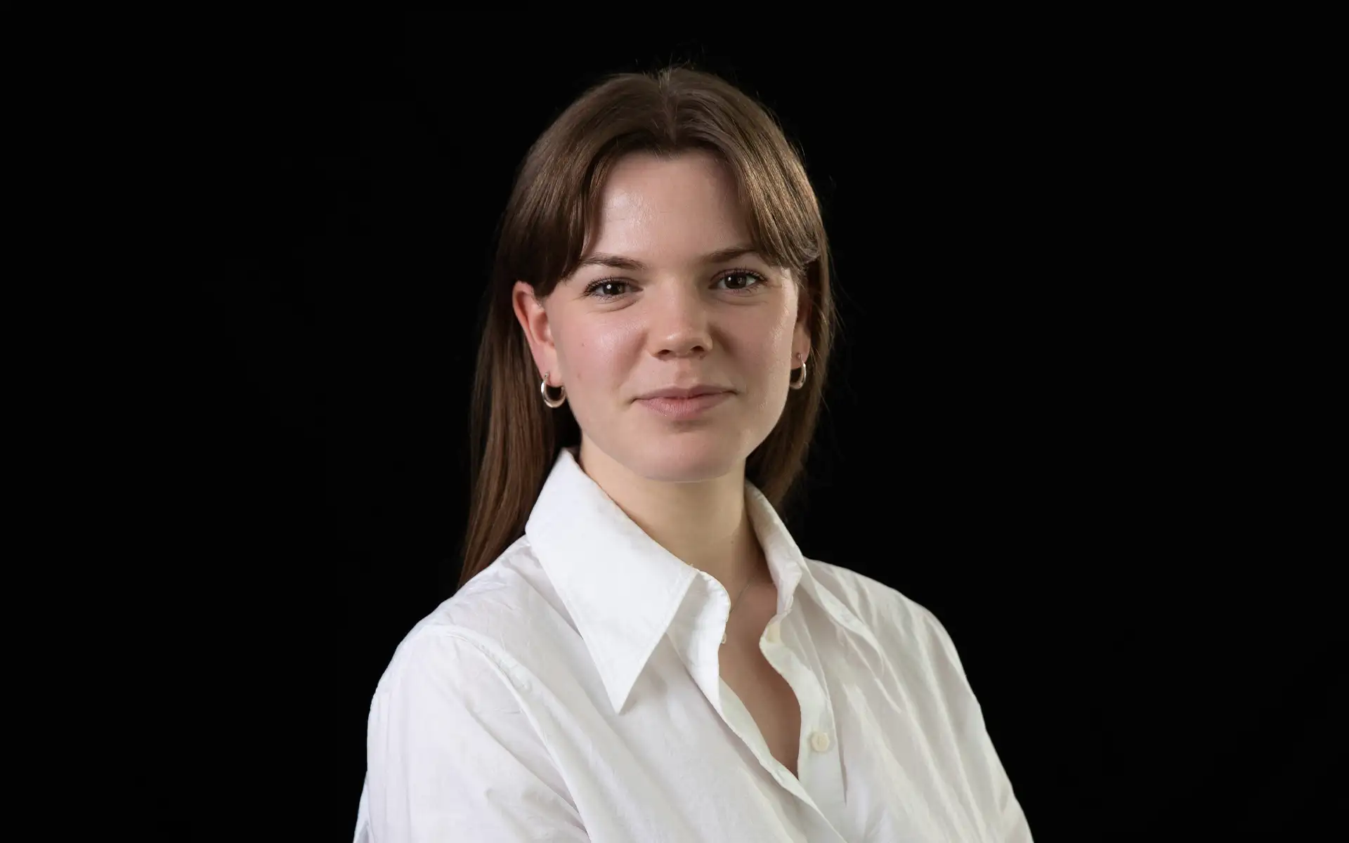 A person with straight, shoulder‑length hair is shown from the chest up against a plain dark background. The person is wearing a white collared shirt and two simple necklaces, one with a small round pendant. Small hoop earrings are visible. The pose is forward‑facing with relaxed shoulders.