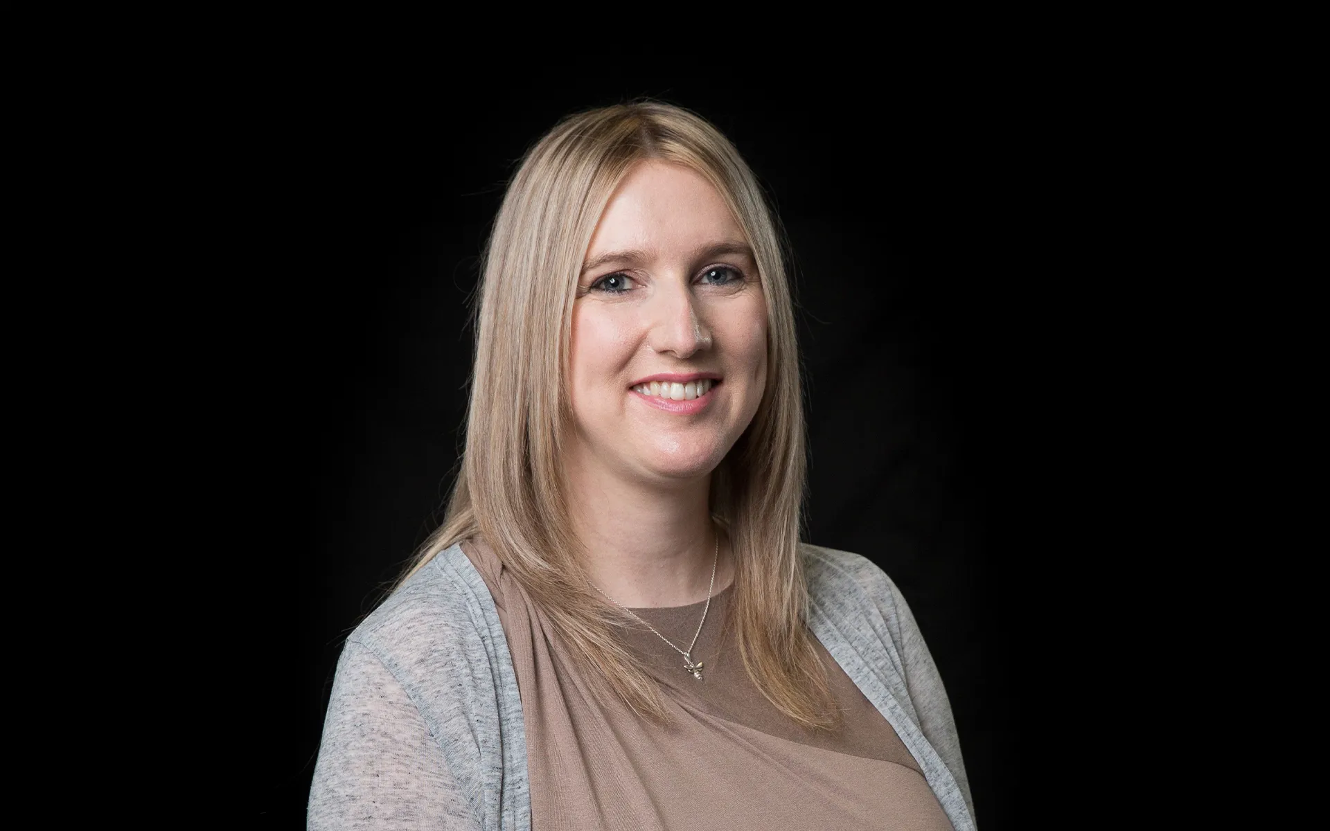 Studio portrait of a person with long light hair, wearing a taupe top and light grey cardigan, photographed against a black background.