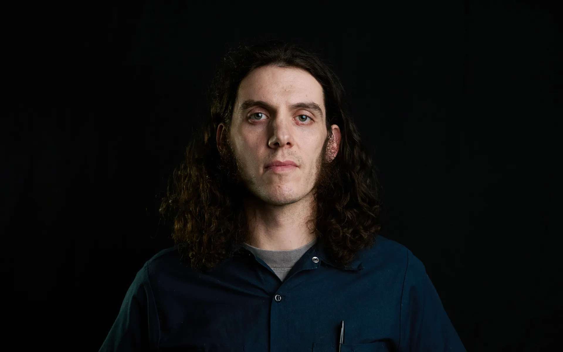 Studio portrait of a person with shoulder-length, curly dark hair, photographed against a black background. The person faces forward with a neutral expression and wears a dark blue button-up shirt with a pen visible in the chest pocket. Soft directional lighting highlights the face while the background remains dark.