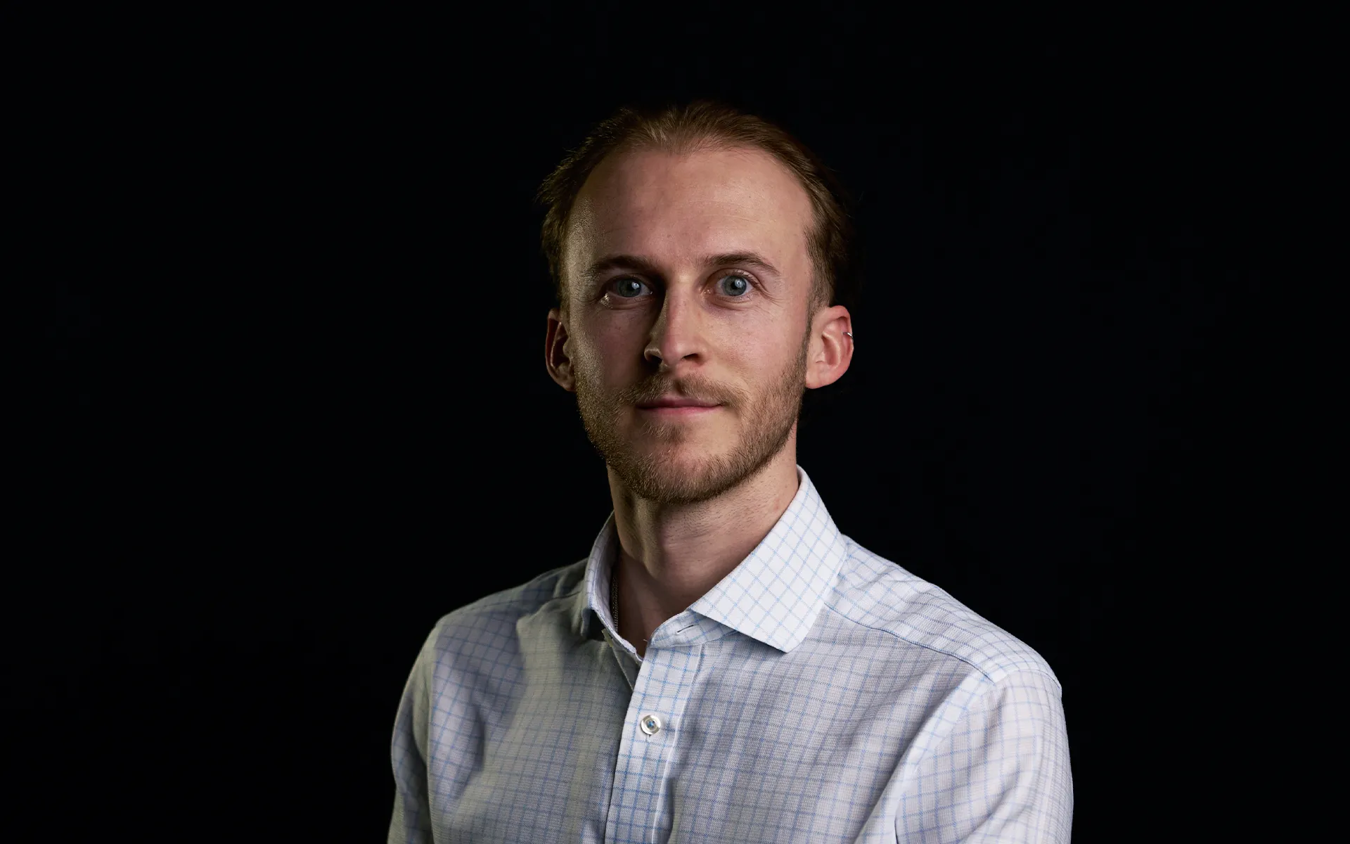 Studio portrait of a person with short hair and light facial hair, photographed against a black background. The person is angled slightly to one side, looking towards the camera, and wears a light-coloured check-pattern shirt. Controlled lighting creates strong contrast between the subject and the dark backdrop.