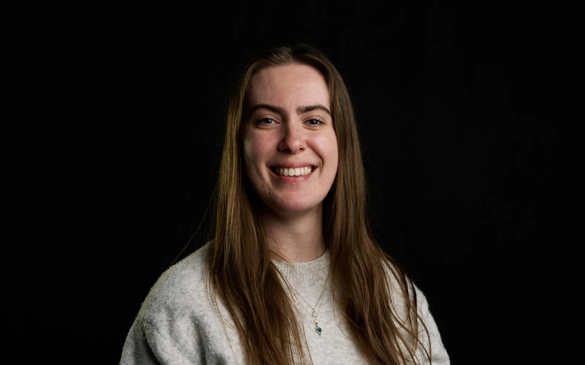 Studio portrait of a person with long straight hair, photographed against a black background. The person is turned slightly to one side, smiling, and wears a light-coloured top with a small pendant necklace. Soft lighting illuminates the face while maintaining a dark, uncluttered background.