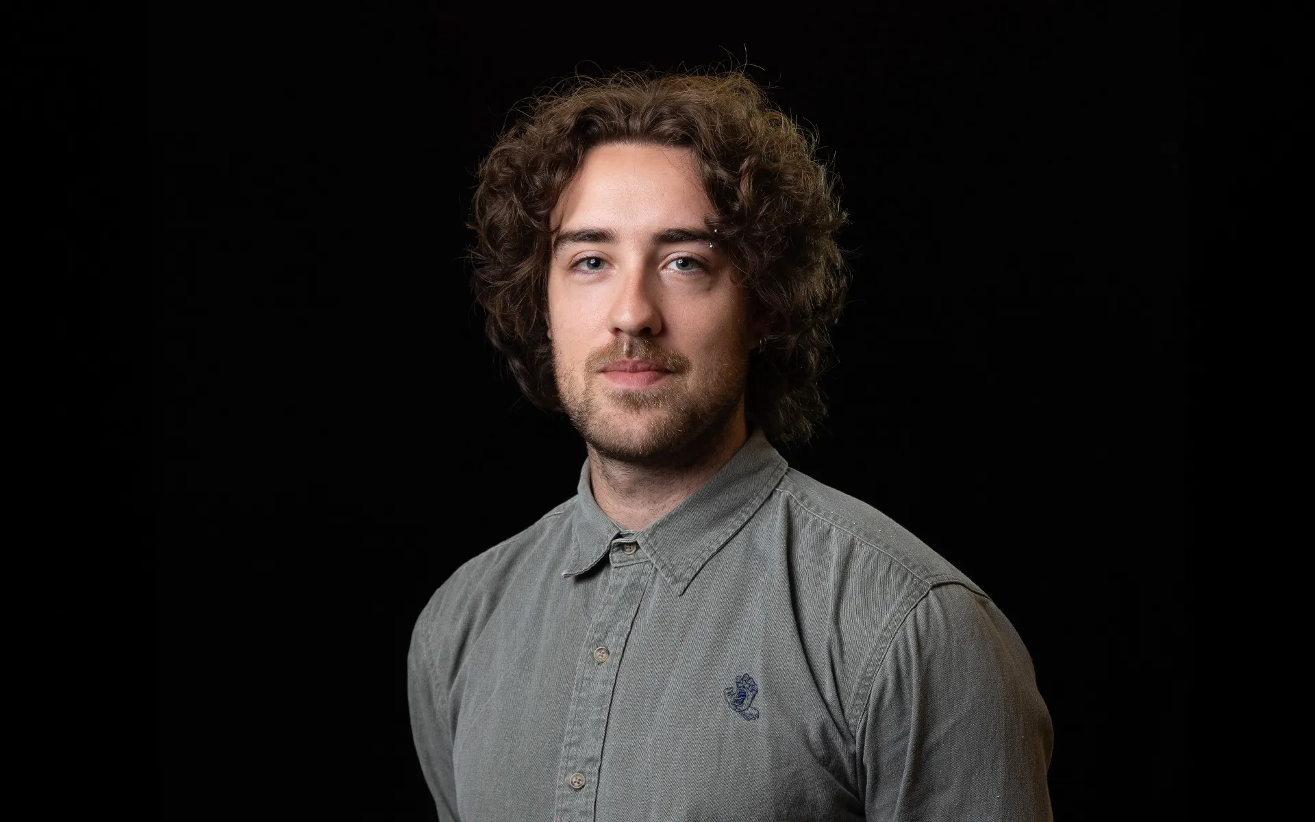 Studio portrait of a person with medium-length curly hair, wearing a grey button-up shirt against a black background.