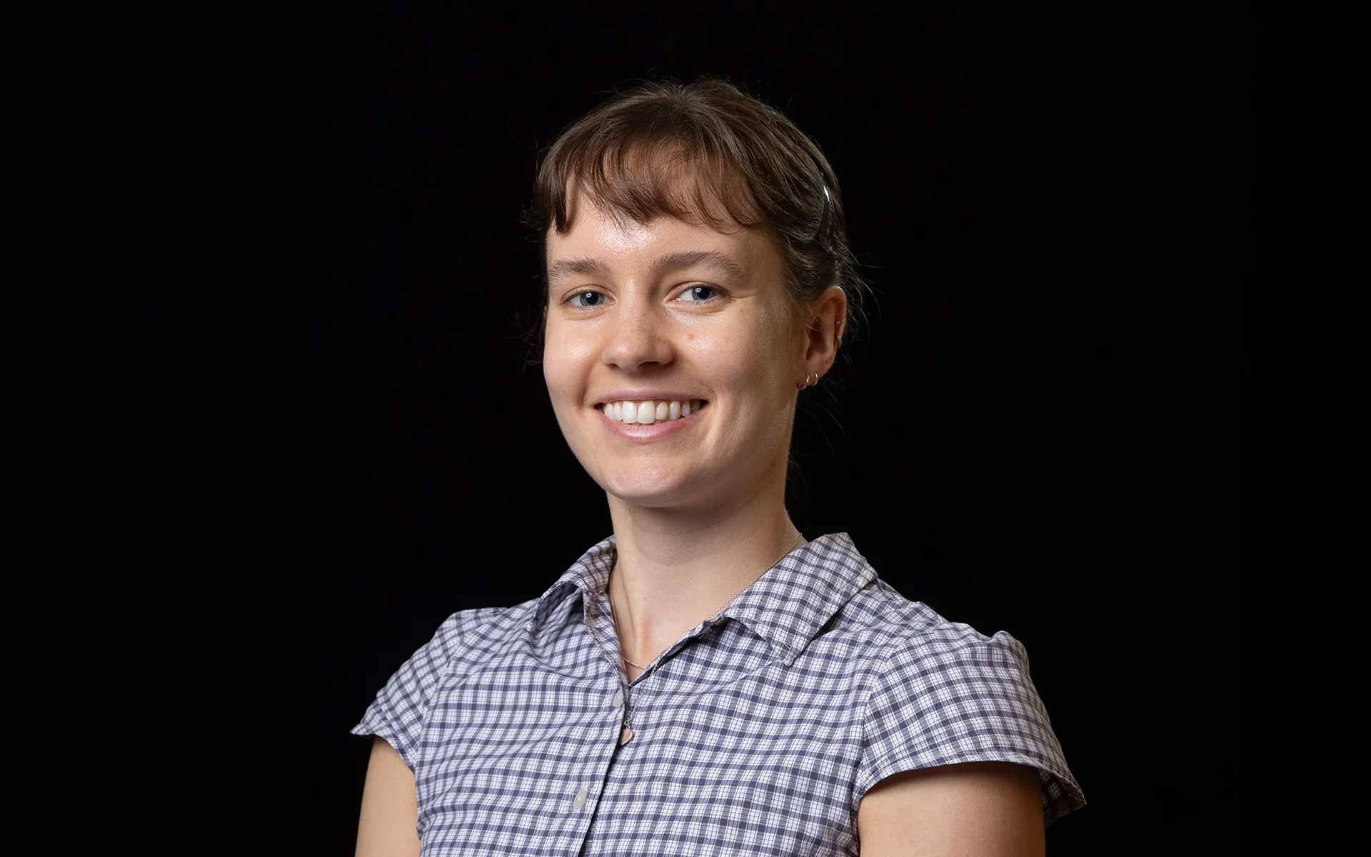 Studio portrait of a person with hair tied back and a fringe, wearing a checked short-sleeved shirt against a black background.