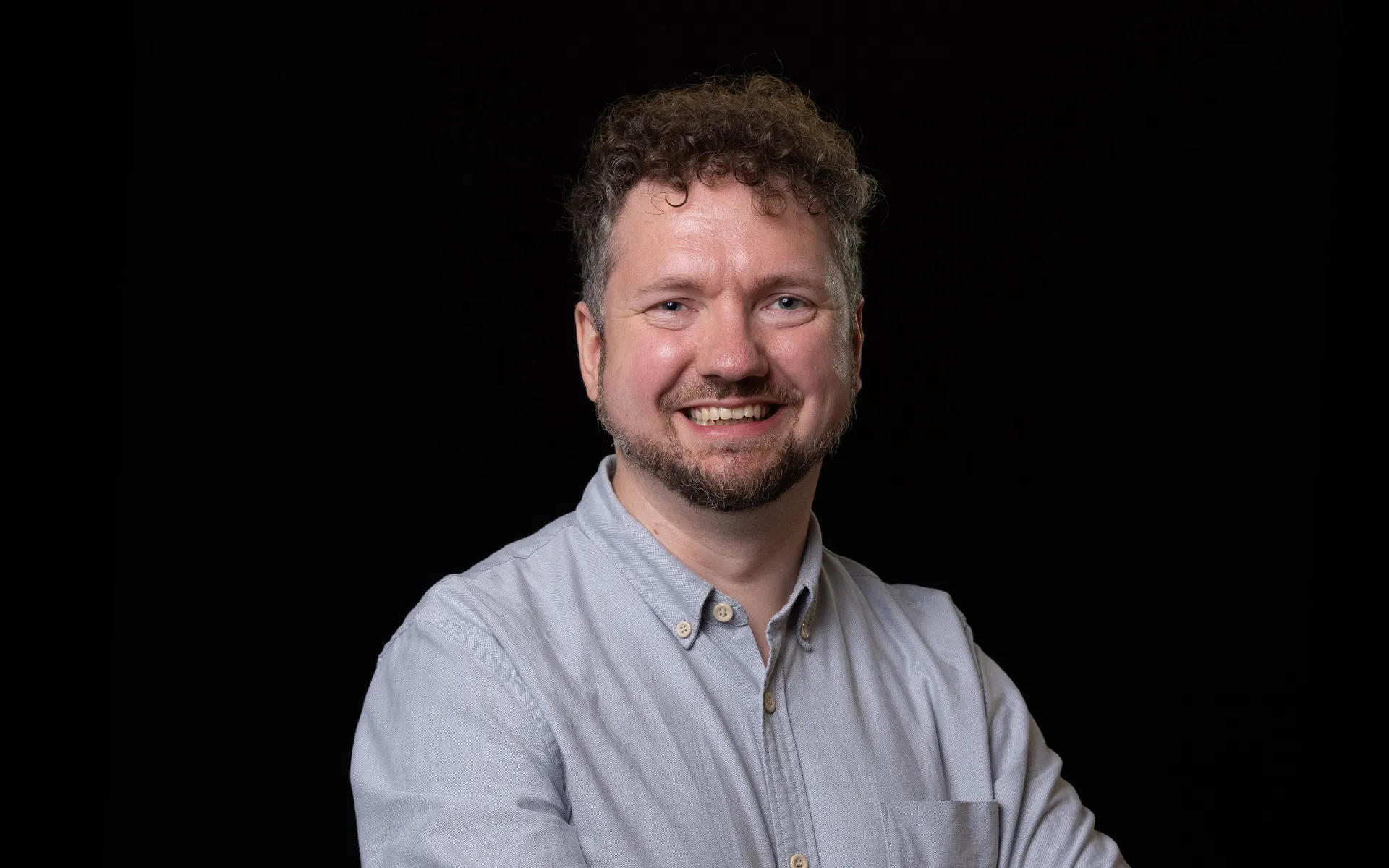 Studio portrait of a person with short curly hair and a beard, wearing a light grey button-up shirt against a black background.