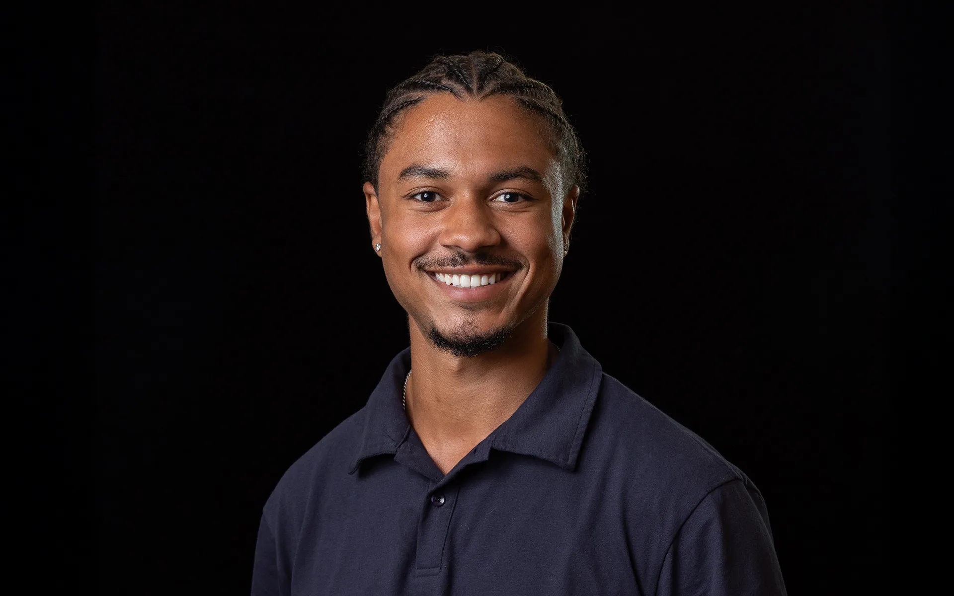 Studio portrait of a person with braided hair, wearing a dark polo shirt against a black background.