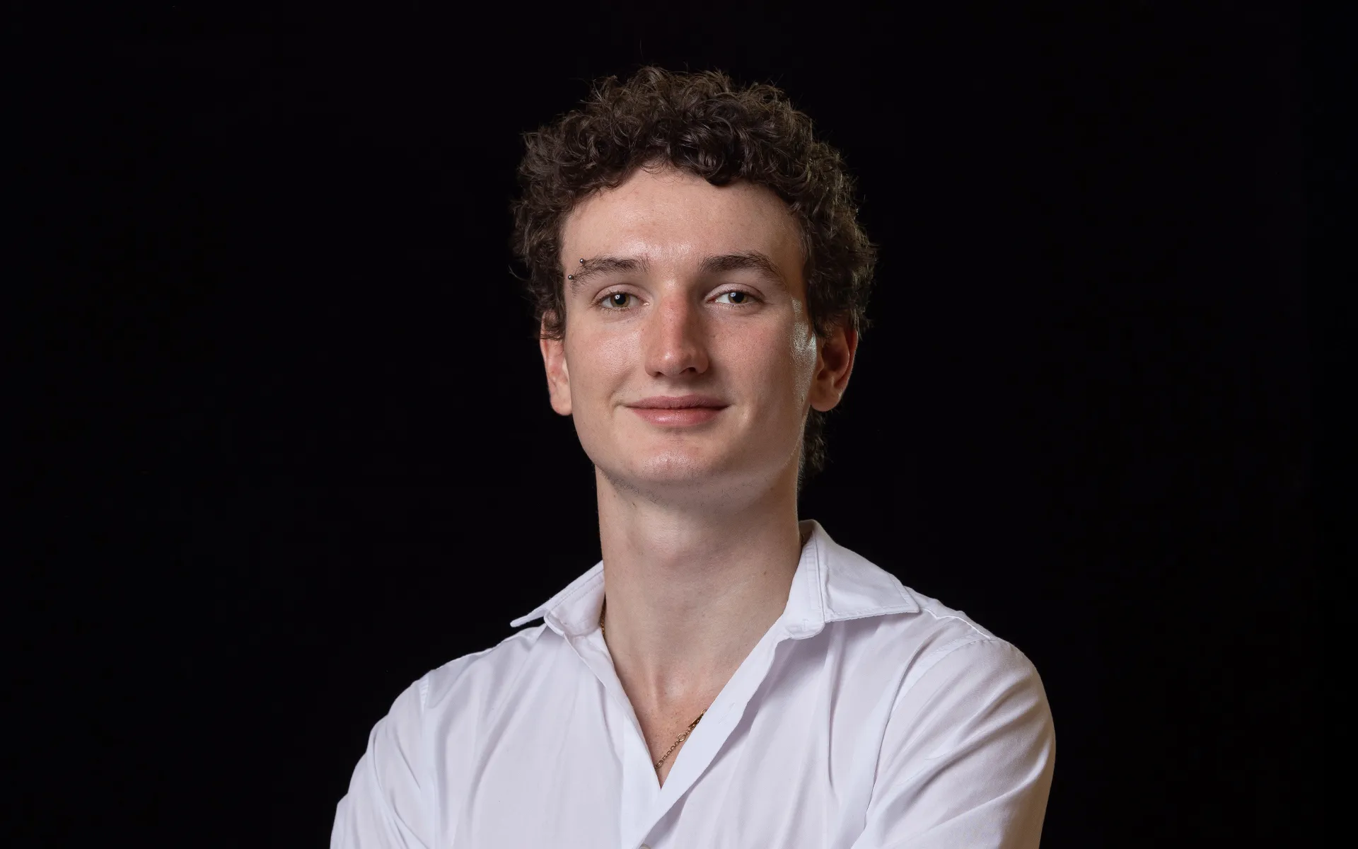 Studio portrait of a person with short curly hair, wearing a white open-collar shirt against a black background.