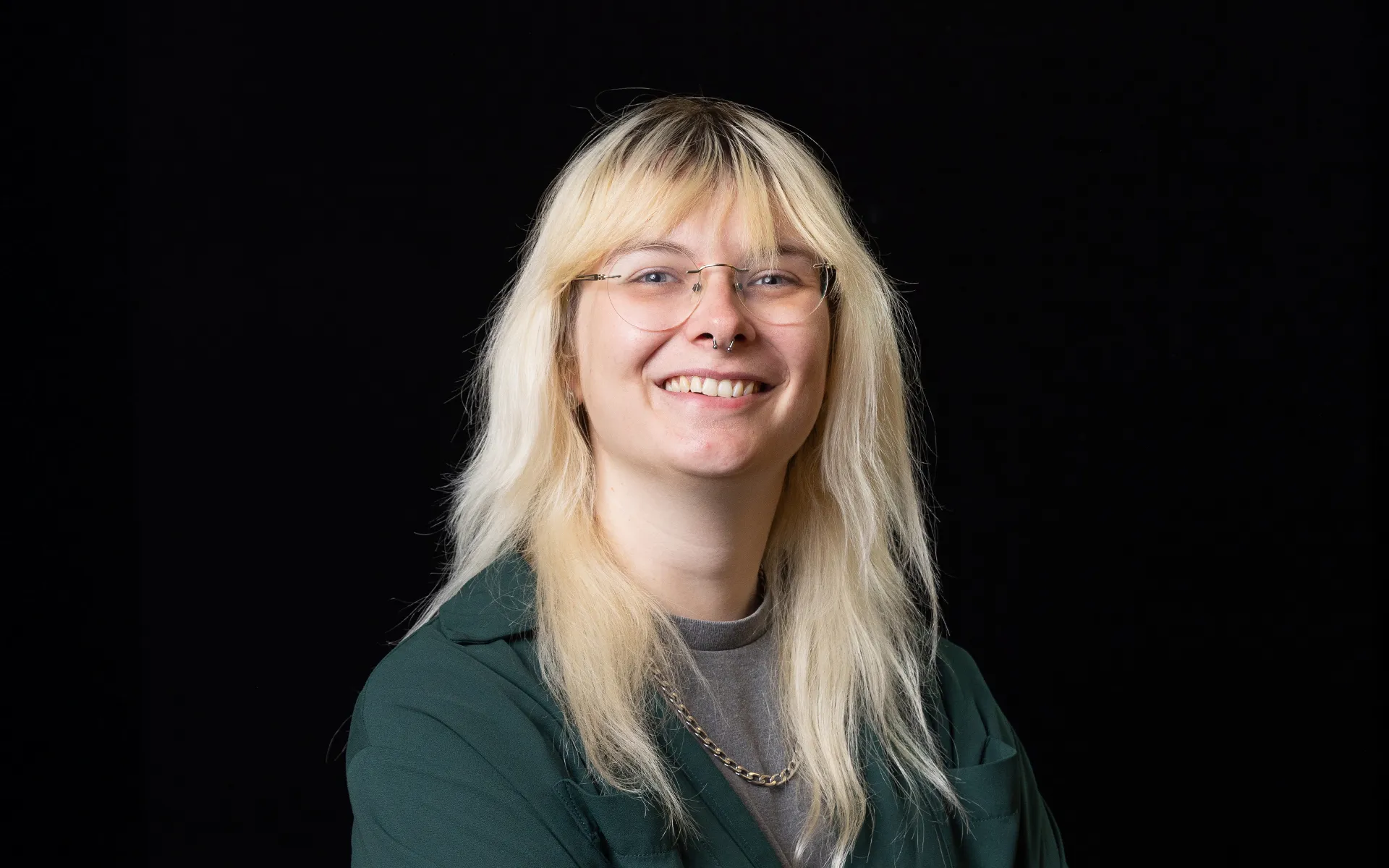 Studio portrait of a person with light-coloured hair and glasses, wearing a green jacket against a black background.