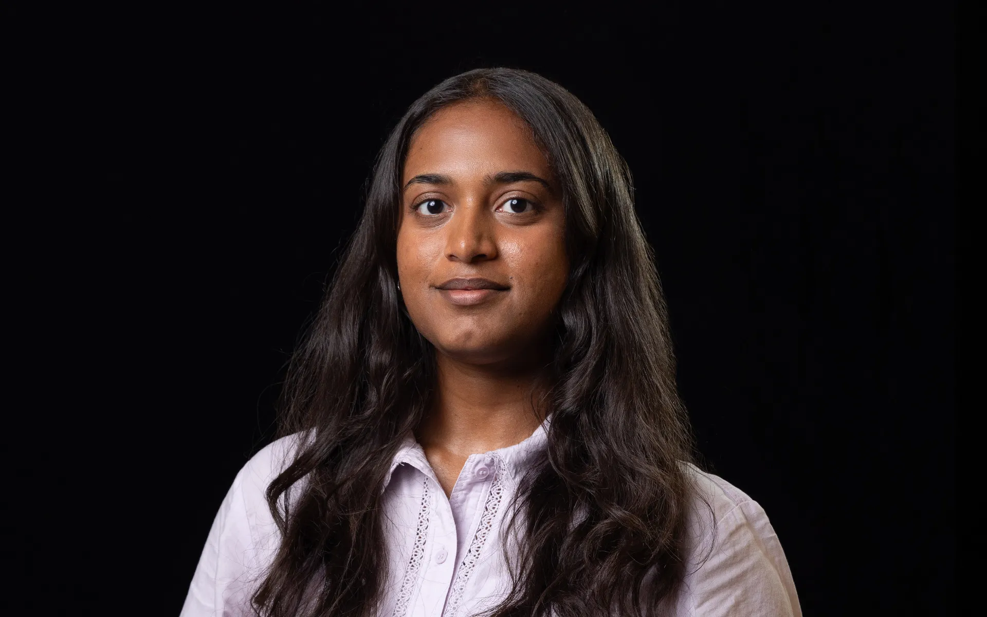 Studio portrait of a person with long dark hair, wearing a pale shirt against a black background.