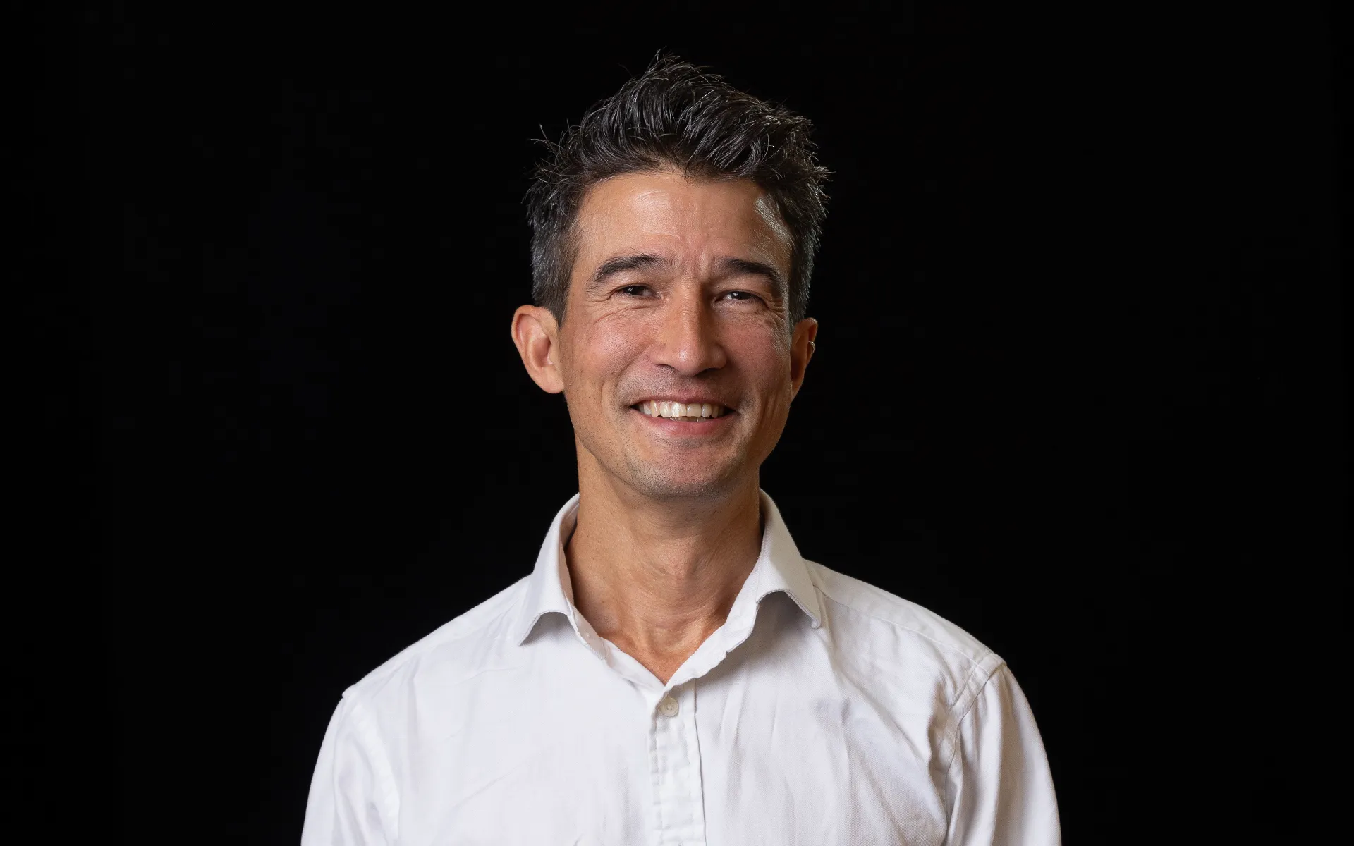 Studio portrait of a person with short dark hair, wearing a white shirt against a black background.