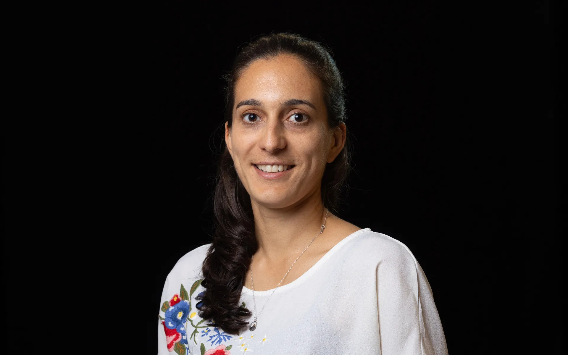 Studio portrait of a person with dark hair styled over one shoulder, wearing a white top with floral embroidery against a black background.