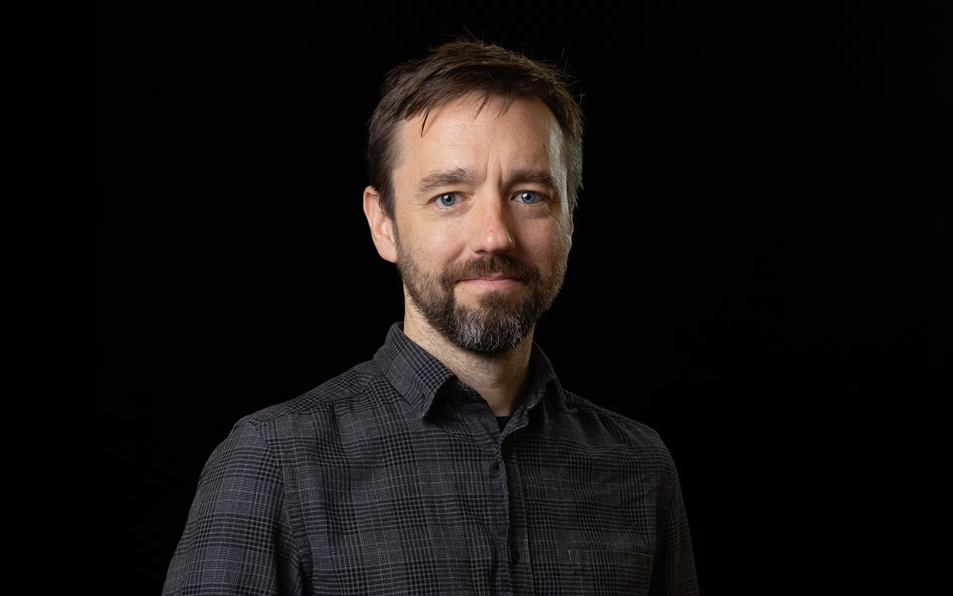 Studio portrait of a person with short hair and a beard, wearing a dark checked shirt against a black background.