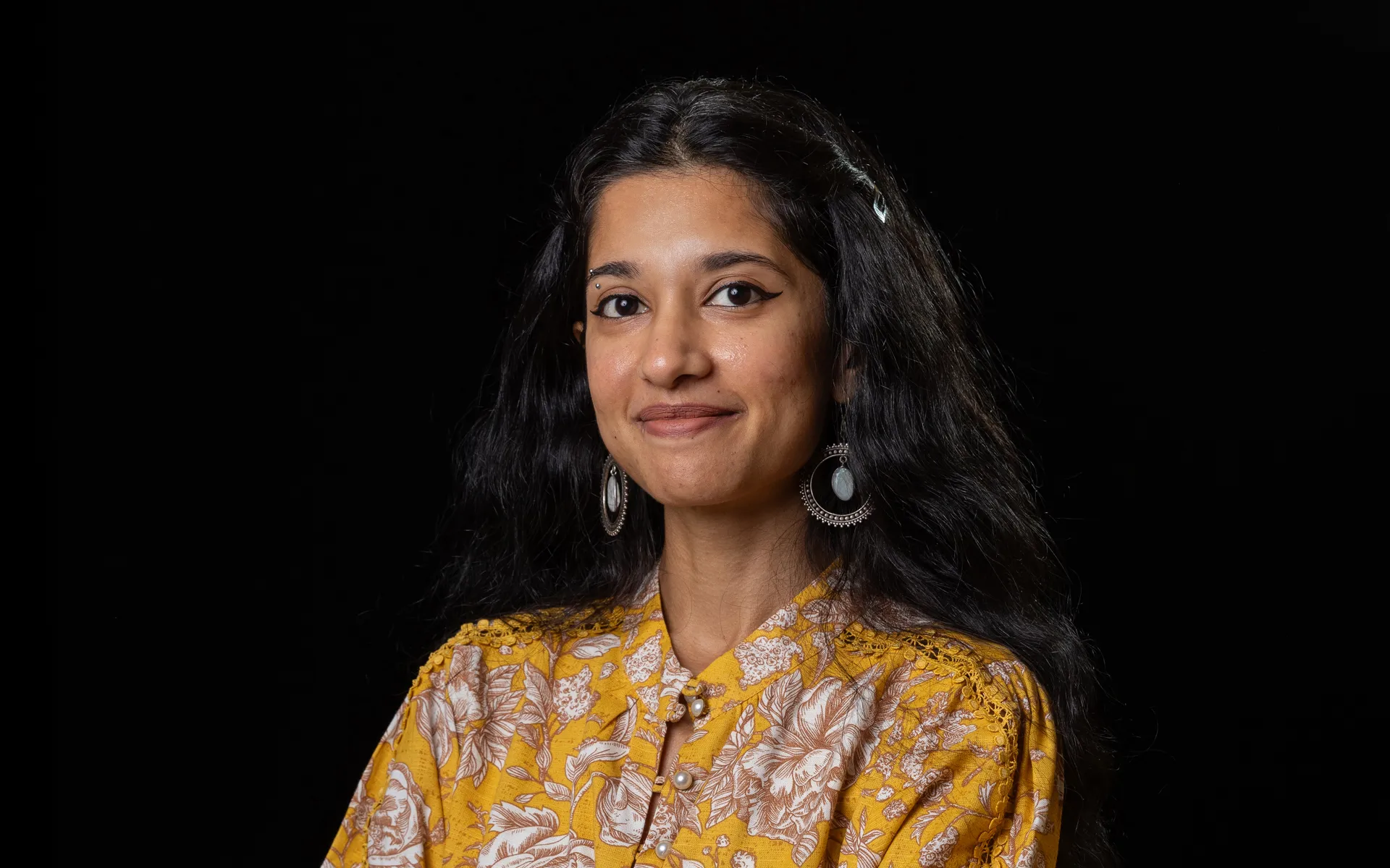 Studio portrait of a person with long dark hair, wearing a yellow patterned blouse and large earrings against a black background.