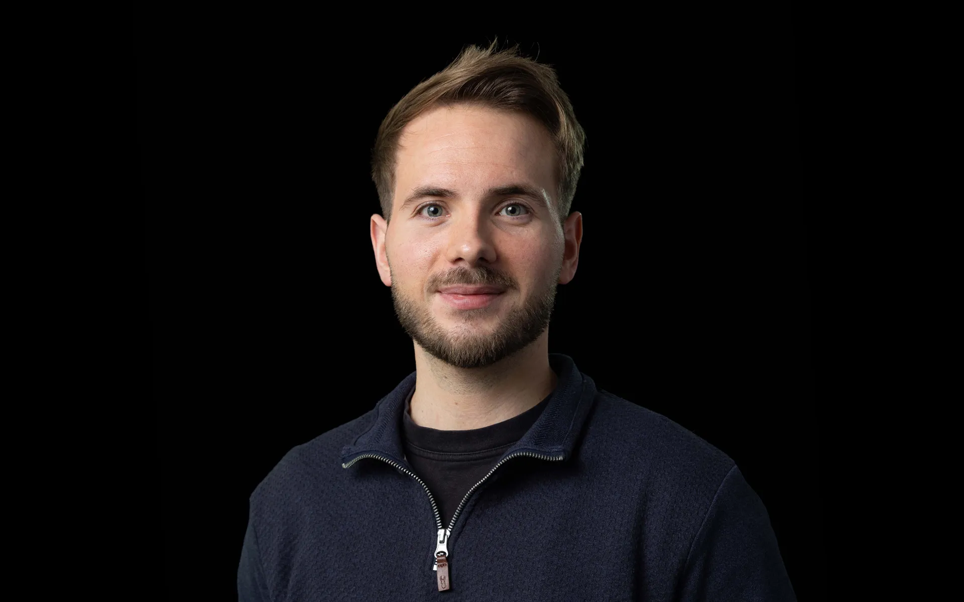 A portrait of Alasdair Moore wearing a dark navy jumper against a black background.