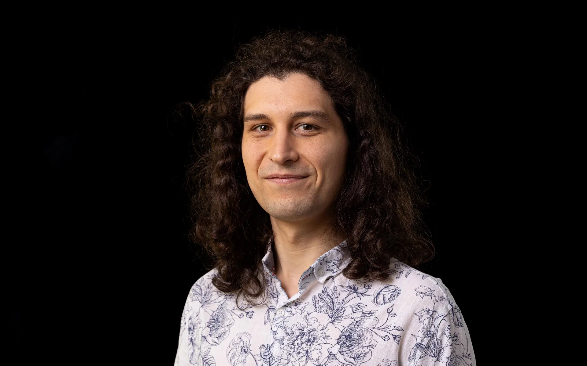 Portrtait of Bartosz Muszkat wearing a white and blue floral shirt against a black background.