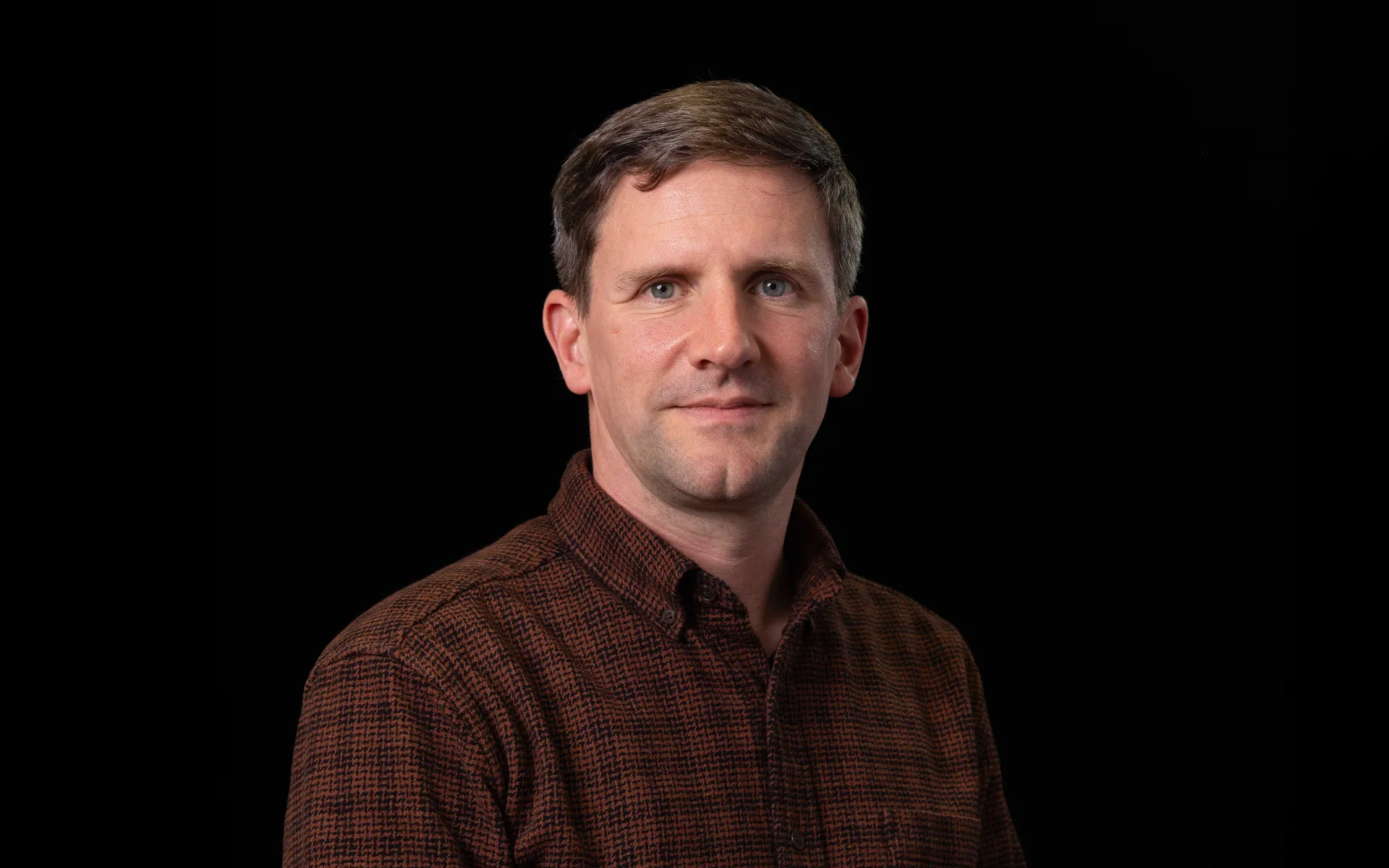 Portrait of Duncan MacLennan wearing a brown shirt against a black background.