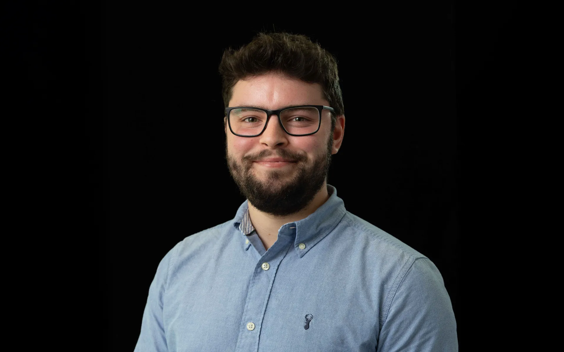 Portrait of Jacob Falconer wearing a light blue button-up shirt and glasses against a black background.