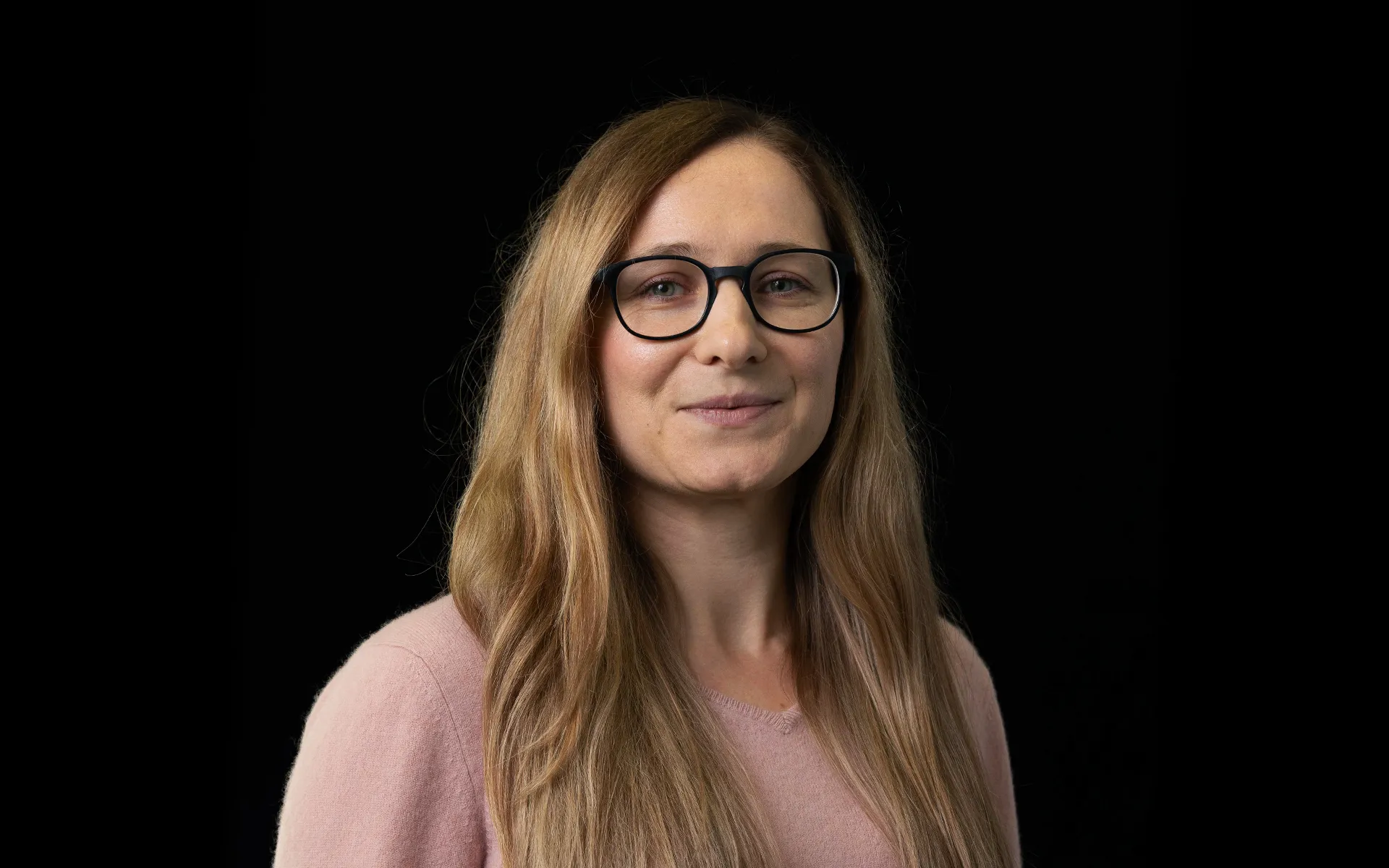Portrait of Kasia Kozlowska wearing glasses and a light pink jumper against a black background.
