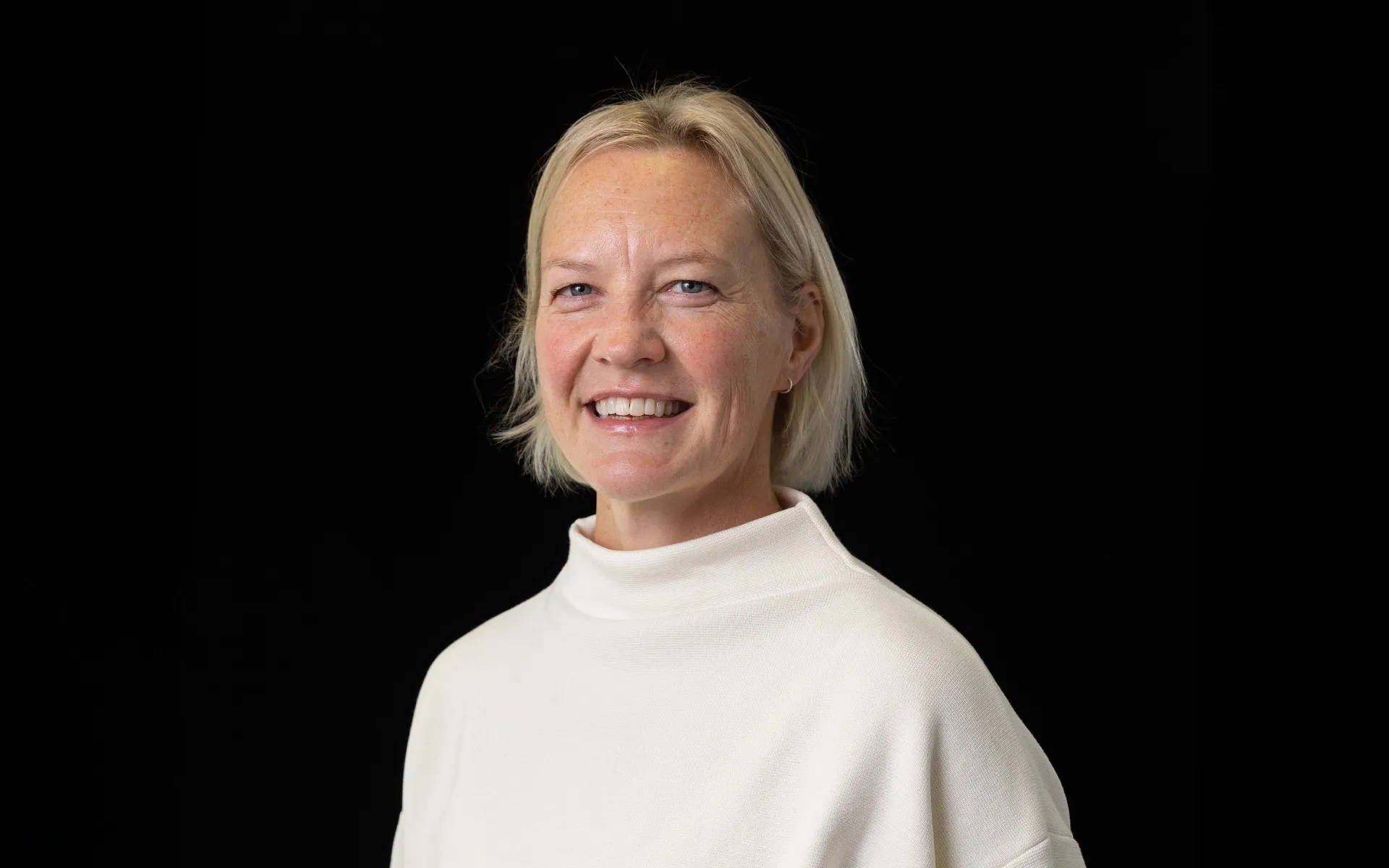 Portrait of Krista Cook wearing a high necked white top against a black background.