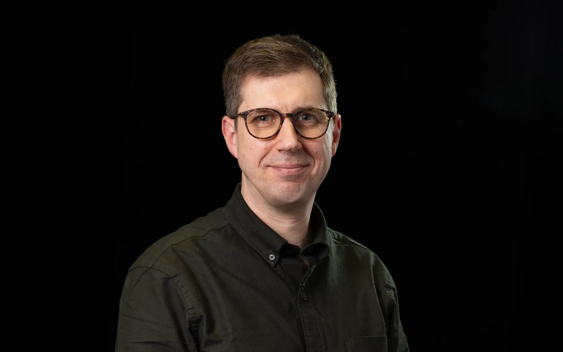 Portrait of Matt Thomas wearing a dark grey shirt and glasses against a black background.