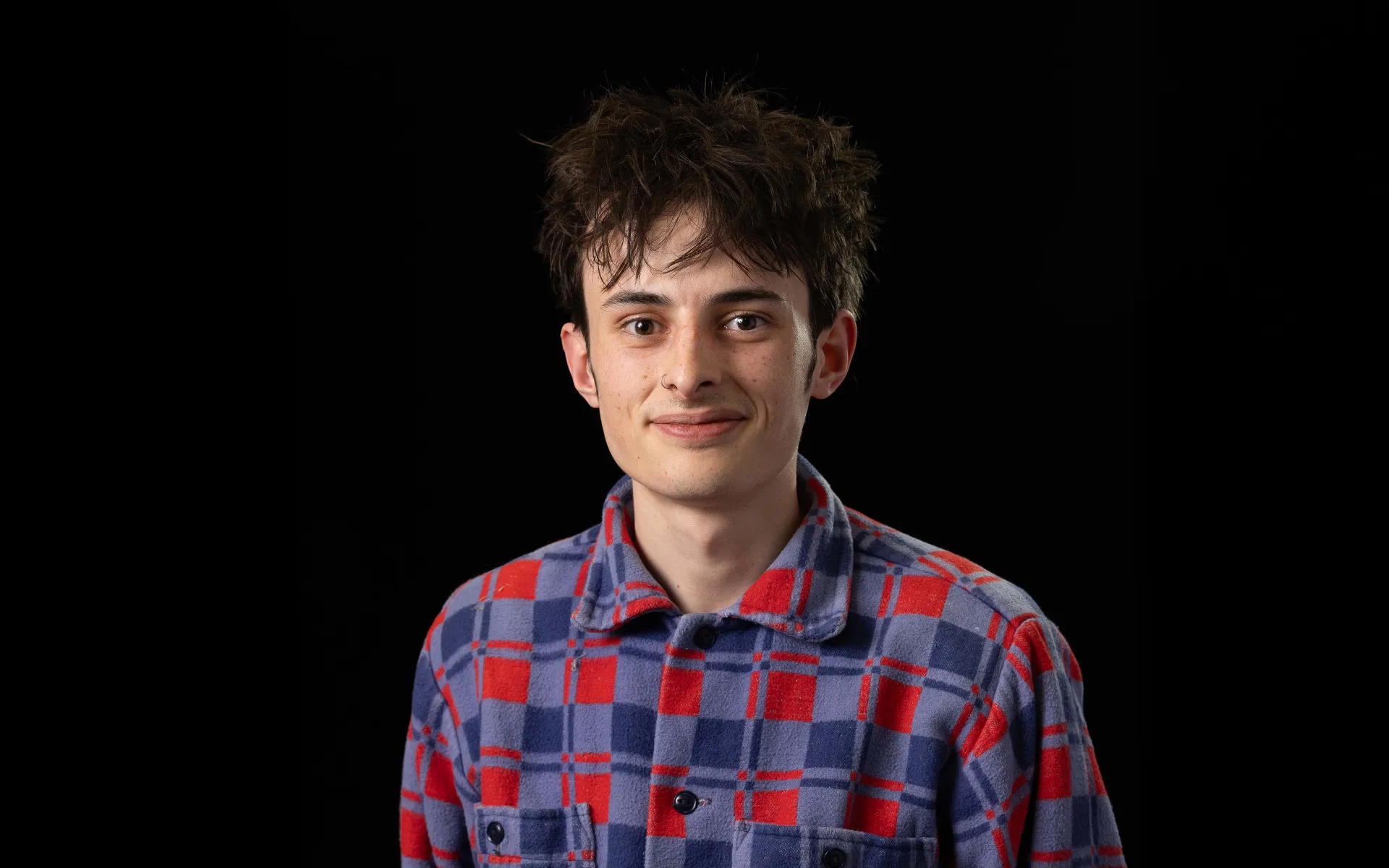Portrait of Paddy Davison wearing a red and blue checked overshirt against a black background.