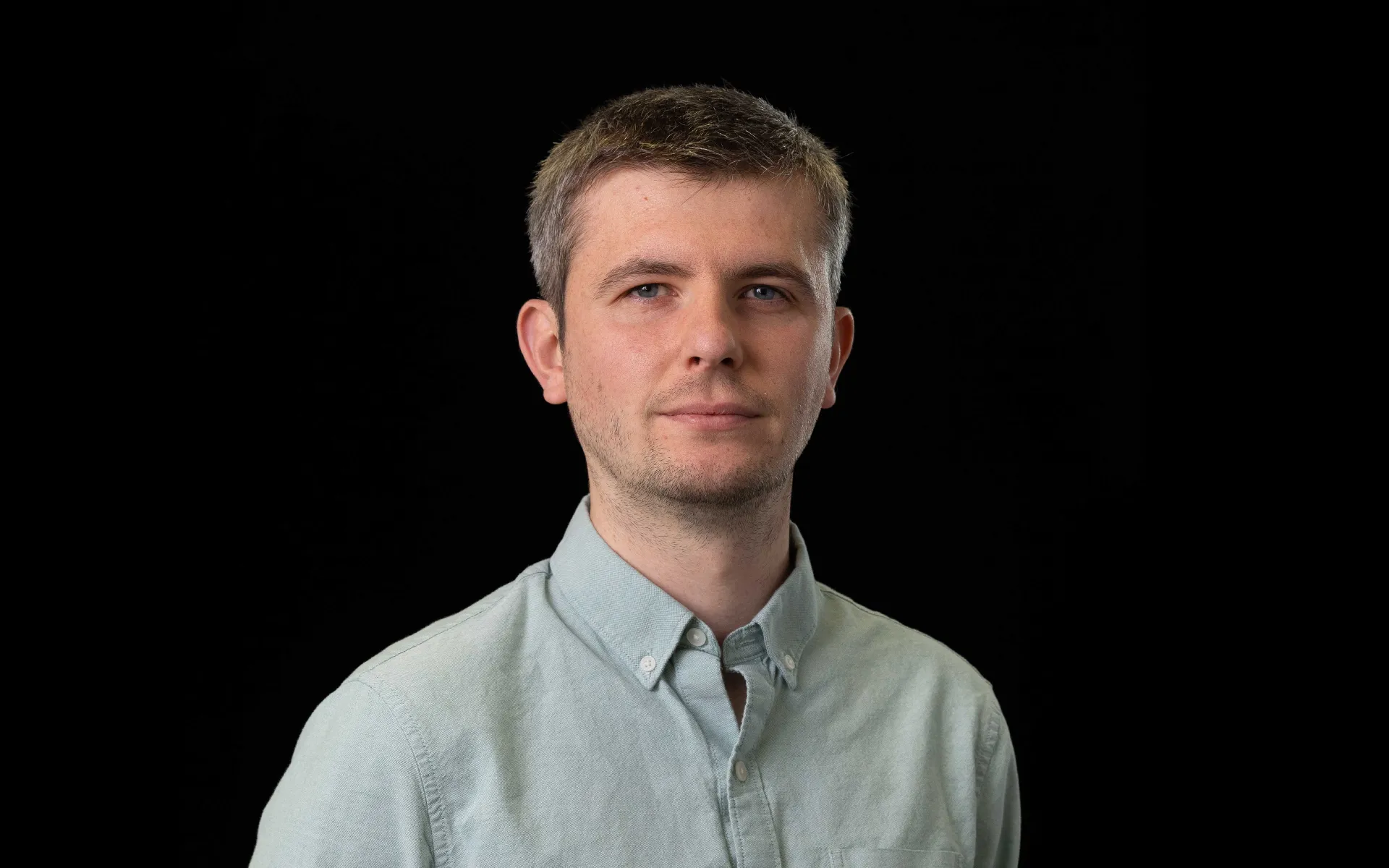 Portrait of Rhys Houston wearing a pale green button-up shirt against a black background.