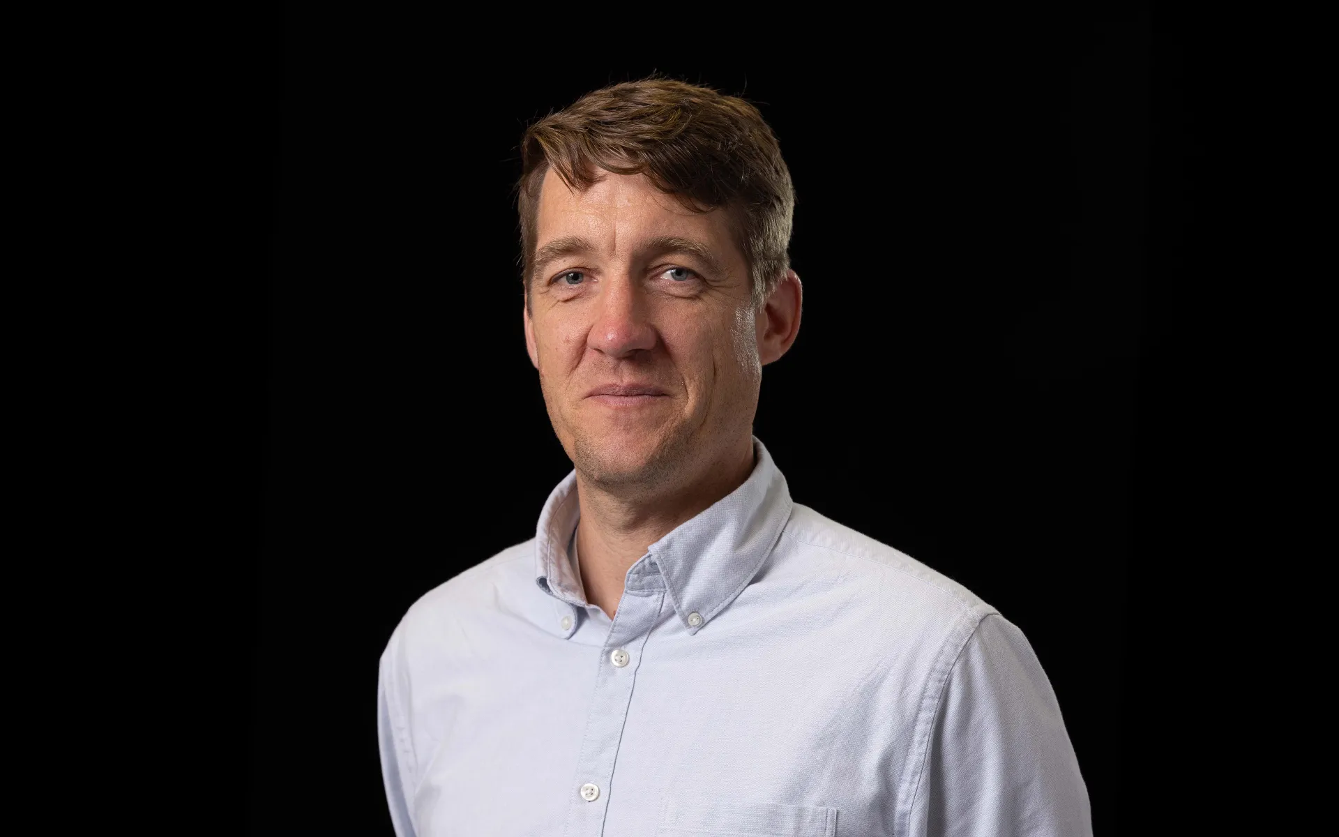 Portrait of Sam Avard wearing a light blue button-up shirt against a black background.