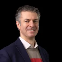 A headshot of Gwilym Still smiling at the camera, wearing a dark jacket and white shirt
