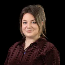 Studio portrait of a woman with hair tied back, wearing a dark patterned blouse and a necklace, photographed against a black background.