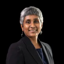 Studio portrait of a woman with short grey hair wearing a dark blazer over a top, photographed against a black background.