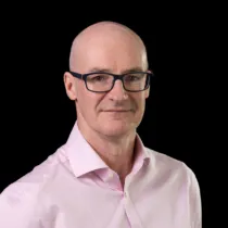 Studio portrait of a man wearing glasses and a light pink shirt, photographed against a black background.