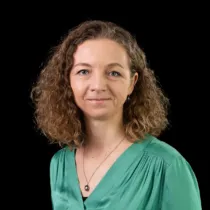 Studio portrait of a woman with curly shoulder-length hair wearing a green blouse and necklace, photographed against a black background.