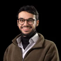 Studio portrait of a person wearing a white collared shirt layered under a dark zip-up fleece, standing against a plain black background.