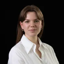 A person with straight, shoulder‑length hair is shown from the chest up against a plain dark background. The person is wearing a white collared shirt and two simple necklaces, one with a small round pendant. Small hoop earrings are visible. The pose is forward‑facing with relaxed shoulders.