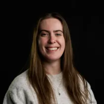 Studio portrait of a person with long straight hair, photographed against a black background. The person is turned slightly to one side, smiling, and wears a light-coloured top with a small pendant necklace. Soft lighting illuminates the face while maintaining a dark, uncluttered background.