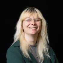 Studio portrait of a person with light-coloured hair and glasses, wearing a green jacket against a black background.