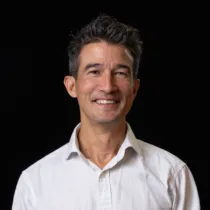 Studio portrait of a person with short dark hair, wearing a white shirt against a black background.