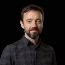 Studio portrait of a person with short hair and a beard, wearing a dark checked shirt against a black background.