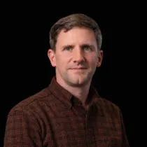 Portrait of Duncan MacLennan wearing a brown shirt against a black background.