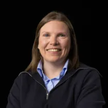 Portrait of Kathryn Donald wearing a light blue shirt under a dark zipped jumper, against a black background.
