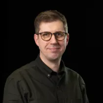 Portrait of Matt Thomas wearing a dark grey shirt and glasses against a black background.