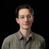 Portrait of Sandy Buchan wearing glasses and an olive-green shirt against a black background.